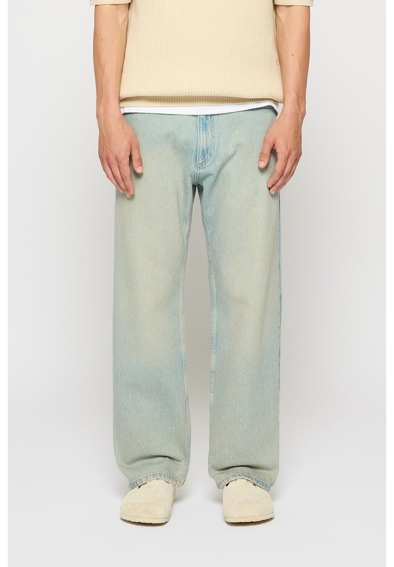 Personne portant un jean large bleu clair, un pull en maille beige et des chaussures crème sans lacets, debout devant un fond uni.