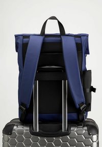 Marinblå ryggsäck med vadderade remmar och nätaccenter, som vilar på en silverfärgad resväska med hexagonmönster, har ett handtag ovanpå och en sidoficka.
