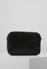 Borsa a tracolla rettangolare in pelle nera e testurizzata con bordi arrotondati, poggiata su un piedistallo bianco su uno sfondo neutro.