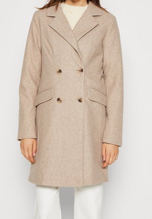 Vrouw draagt een beige wollen mantel met dubbele rij knopen, reverskraag, klepzakken en knopen, gecombineerd met een witte broek en een crèmekleurige trui eronder.