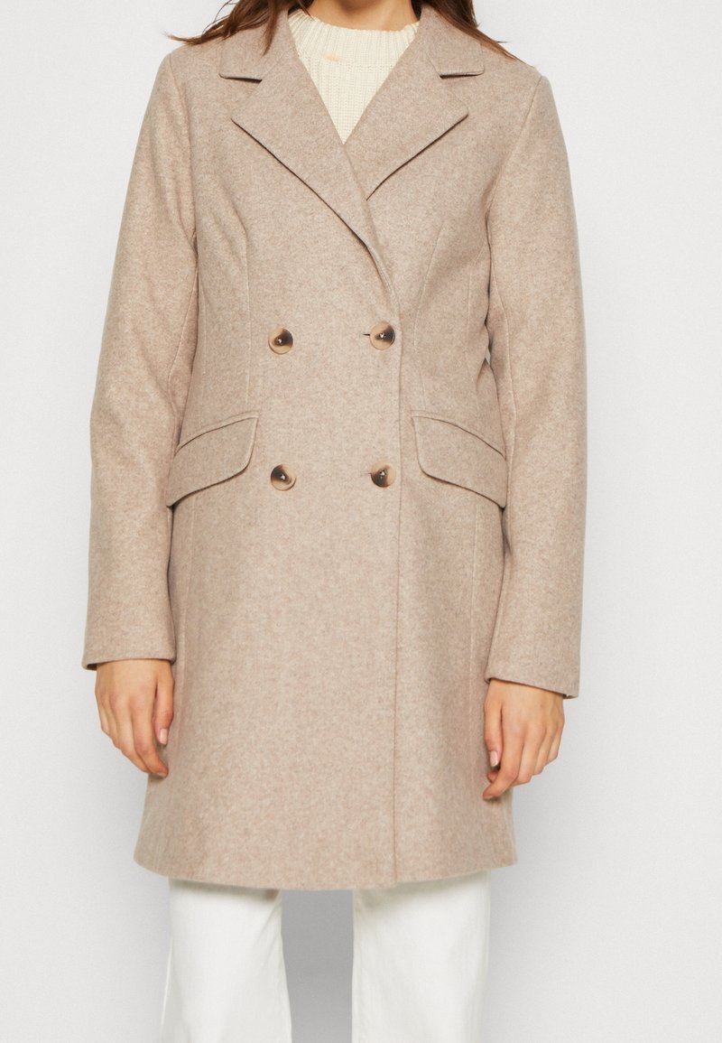 Femme portant un manteau en laine beige à double boutonnage avec revers, poches à rabat et boutons, associé à un pantalon blanc et un pull crème en dessous.