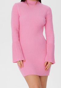 Mini-robe rose côtelée avec manches longues évasées et col montant, réalisée en matière douce en maille, présentant une coupe ajustée et un tissu texturé.