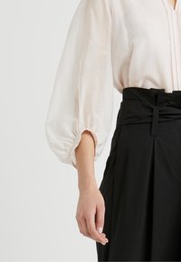 Blouse rose clair, ample, avec des manches évasées et un plastron boutonné, associée à un pantalon noir plissé taille haute.