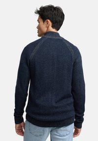 Granatowy sweter z dzianiny o teksturowanym wykończeniu, z prążkowanymi mankietami i dołem oraz rękawami typu raglan. Posiada wysoki kołnierz i luźny krój.