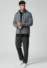 Veste imperméable grise avec un col haut et une poche poitrine zippée, associée à un pantalon noir et des baskets blanches. Matériau lisse et léger.