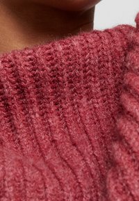 Pull en tricot côtelé de couleur rose profond, offrant une texture douce et un col montant, avec des coutures visibles créant un effet superposé.