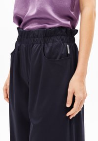 Marineblaue, weiche Stoffhosen mit elastischem Bund, seitlichen Taschen und lockerer Passform. Glatte Textur mit subtilen Stickdetails.