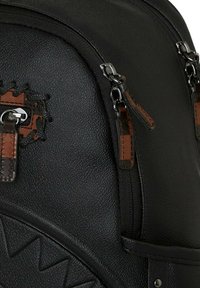 Mochila negra texturizada con acentos y cremalleras de cuero marrón, con un parche decorativo y un detalle de patrón triangular en el costado.