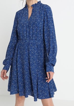 Frau trägt ein langärmliges blaues Kleid mit kleinem weißem Blumenmuster, Rüschenkragen und taillierter Taille, steht mit entspannten Armen.