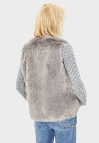 Grå fuskpälsväst med en mjuk textur, ärmlös design och en bakvy; matchad med en grå stickad tröja och blå denimjeans.