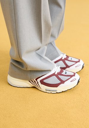 Grijze broek over witte en bordeauxrode Nike-sneakers, staand op een mosterdgele vloerbedekking.