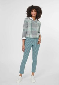 Grijze geblokte trui met groene accenten, gecombineerd met teal geklede broeken met een patroon. Model draagt witte sneakers; casual, getailleerde silhouet.