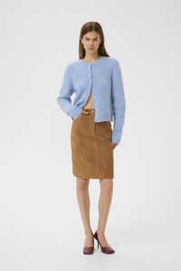 Kvinne står iført en lys blå, puffete cardigan, brun kne-langs skjørt, og mørkerøde høyhælte sko mot en hvit bakgrunn.