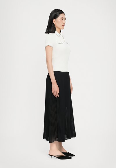 Weißes geripptes Poloshirt mit Kragen und Knopfakzenten, kombiniert mit einem schwarzen plissierten Midirock und schwarzen spitzen Slingback-Heels.