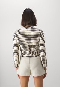 Kobieta z prostymi, ciemnymi włosami do ramion, ubrana w paskowany sweter z długim rękawem i białe szorty z wysokim stanem o fakturowanej powierzchni, stojąca tyłem.