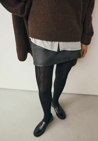 Brauner, übergroßer Strick-Pullover über einem weißen gestreiften Hemd, schwarzem Leder-Minirock, schwarzen Spitzenstrumpfhosen und schwarzen Loafers.
