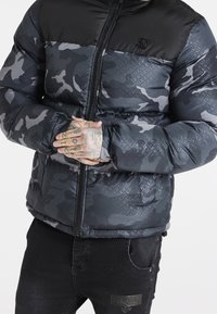 Giacca imbottita nera e grigia con motivo mimetico e dettagli a marchio, dotata di chiusura con zip completa e polsini elasticizzati.