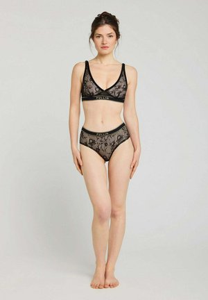Femme debout pieds nus portant un bralette en dentelle noire et une culotte taille haute avec le logo de la marque Pullin sur les bandes élastiques, sur un fond clair uni.