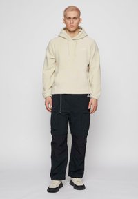 Beige fleece hoodie met voorzak en trekkoorden, gedragen met zwarte cargo pants met meerdere zakken en verstelbare banden. Lichte sneakers.