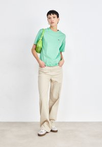 Camiseta de algodón verde claro con cuello redondo, combinada con pantalones de pierna ancha beige y zapatillas plateadas. Accesorios con un pequeño bolso verde.