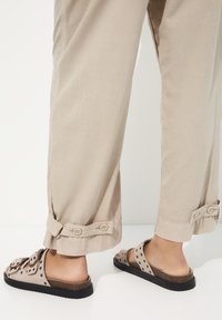Pantalons beige amples avec poignets roulés, dotés de deux boutons. Associés à des sandales à glissière brunes avec des découpes et une semelle noire.