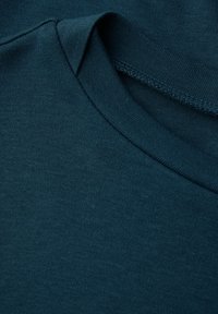 Teal jersey-kangas pyöreällä kauluksella ja ribbireunuksilla. Tekstuuri on sileä, hienovaraisella kiillolla ja näkyvät ompeluyksityiskohdat ovat vähäiset.