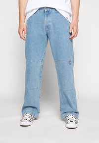Lys blå denimbukser med en avslappet passform, med knelapper og et logo-patch på siden. Stylet med svart-hvite mønstrede sko.