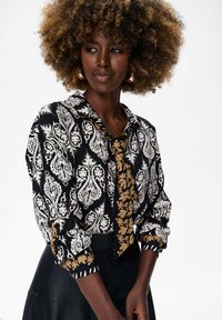 Vrouw met krullend haar die een gestreepte zwart-witte blouse met gouden bloemenaccenten en een zwarte rok draagt, en iets opzij kijkt.