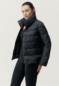 Črna puhovka z visokim ovratnikom, s prešitim dizajnom, s pritrdilnimi zapirali in gladkim, sijajnim zaključkom. Nosila jo je čez črne pajkice.