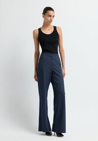 Vrouw in een zwarte mouwloze top, marineblauwe streepjes brede broek en zwarte hoge hakken die tegen een effen lichte achtergrond staat.