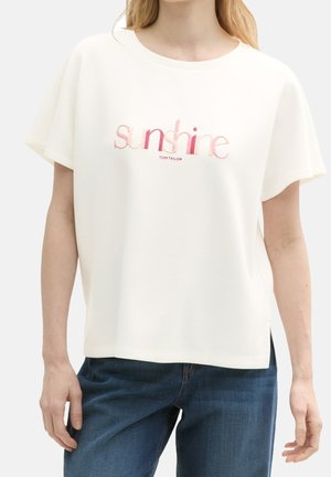 Frau trägt ein weißes T-Shirt mit kurzen Ärmeln, auf dem in pinken Buchstaben "Sunshine" steht, und blaue Jeans vor einem einfarbigen Hintergrund.