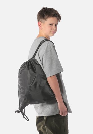 Ragazzo con maglietta grigia e pantaloni verdi guarda oltre la spalla portando uno zaino nero con coulisse su sfondo bianco.