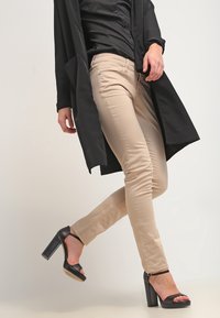 Beiga skinny jeans med en mjuk textur, kombinerat med en svart lång kappa och svarta plattformssandaler, vilket lyfter fram outfitens lagerdesign.