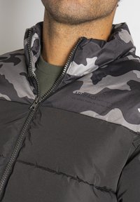 Schwarze gepolsterte Jacke mit einem grauen Camouflage-Muster am Kragen. Verfügt über einen Reißverschluss vorne und eine glatte Textur. Markenname sichtbar.