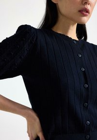 Cardigan lavorato a maglia blu navy con texture a coste, maniche a palloncino corte e chiusura con sei bottoni. Presenta un collo scallop e un tessuto morbido.