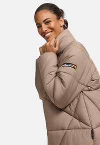 Beige pufferjack met een hoge kraag, een ruitvormig quiltpatroon en een kleurrijke logopatch op de linker mouw. Zachte, gewatteerde textuur.
