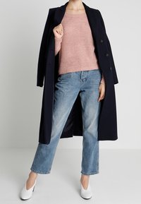 Eine Person trägt einen dunkelblauen Mantel über den Schultern, einen rosa Strickpullover, hellblaue Jeans und weiße Absatzschuhe und steht vor einem einfarbigen Hintergrund.