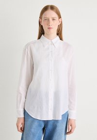 White long-sleeve shirt made of lightweight fabric, featuring a button-up front, collar, and a curved hemline. Minimalist design and soft texture.