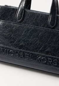 Sac à main en cuir noir texturé avec deux poignées solides. Présente le logo "MICHAEL KORS" en relief sur une section lisse. La finition craquelée ajoute du détail.