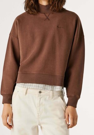 Persona que lleva una sudadera corta marrón sobre una camisa de cuadros, combinada con unos pantalones de talle alto color blanco roto, de pie frente a un fondo liso.