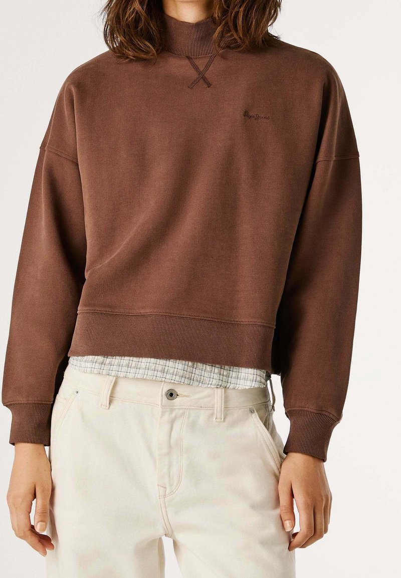 Personne portant un sweat court marron sur une chemise à carreaux, associé à un pantalon taille haute écru, debout devant un fond uni.