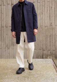 Marineblauer Mantel mit Knopfverschluss, einer Brusttasche, getragen über einem schwarzen Oberteil und cremefarbenen Hosen, kombiniert mit schwarzen Turnschuhen.