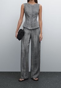 Graues Leinen-Top ohne Ärmel, kombiniert mit weit geschnittenen grauen Hosen. Der Stoff hat eine strukturierte Oberfläche. In einer Hand wird eine schwarze Clutch gehalten.