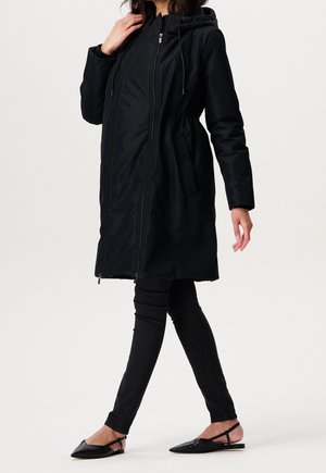 Parka negra con capucha, cierre frontal con cremallera, cintura ajustada, dos bolsillos laterales y mangas largas, combinada con jeans skinny negros y bailarinas.