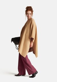 Béžový vlnený pončo s širokými rukávy a károvanou podšívkou. Model nosí bordové kalhoty s širokými nohavicemi a černé kotníkové boty.