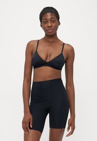 Slank kvinne med kort hår iført svart BH og høyt liv svarte sykkelshorts, stående mot en enkel hvit bakgrunn.