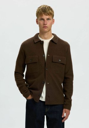 Jeune homme portant une veste en laine marron foncé avec des poches poitrine boutonnées, par-dessus une chemise blanche et un jean foncé, debout devant un fond uni.