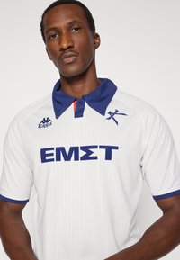 Biele polo tričko s krátkym rukávom, modrým golierom a manžetami, s výraznou modrou grafikou "EMΣT" na prednej strane a logom Kappa na ľavej strane hrudníka.