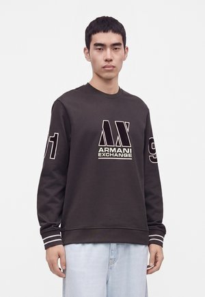 Młody mężczyzna noszący czarną bluzę Armani Exchange z dużym logo i liczbami na rękawach, w połączeniu z jasnoniebieskimi jeansami, stojący na białym tle.