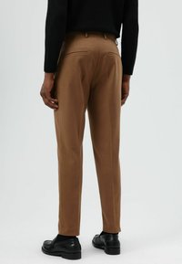 Bruine op maat gemaakte broek met een gladde textuur, voorzien van twee achterzakken, gedragen met zwarte loafers en een zwarte trui.